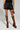 Wearing the BACK TO THE FUTURE MINI SKIRT paired with leopard print knee-high boots featuring platform heels, they project a bold style. A skull tattoo decorates their thigh as they carry a leopard print purse, completing an edgy ensemble. The plain background emphasizes their unique fashion sense.