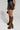A person wearing the BACK TO THE FUTURE MINI SKIRT, a stylish windbreaker-inspired black leather mini skirt, is holding a small bag and wearing leopard print knee-high boots with high heels, standing against a plain background.