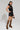 A person poses confidently in the BACK TO THE FUTURE MINI SKIRT made of black faux leather, paired with a matching vest. They sport leopard print platform boots and carry a small matching handbag. The stylish, windbreaker-inspired ensemble stands out against the plain white background, highlighting the outfit's edgy vibe.