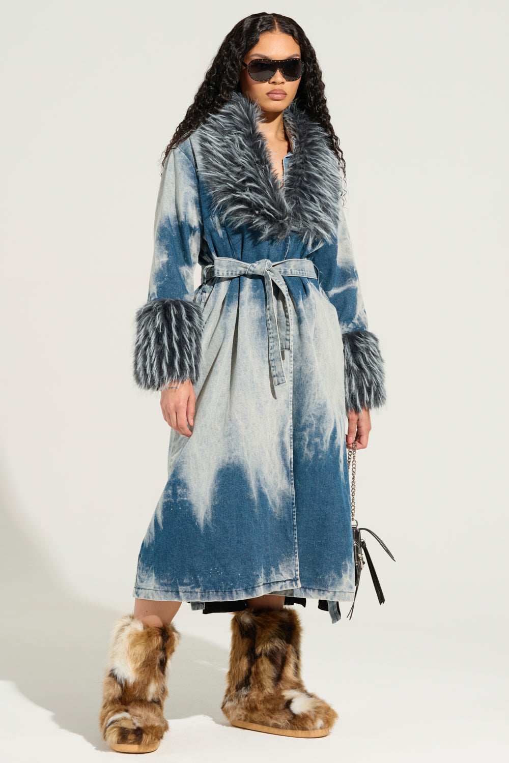 Dressed in the LIV FUR TRIM DENIM TRENCH featuring a luxurious fur-trim collar and cuffs, a person confidently strikes a pose against a plain background. Oversized sunglasses complement the ensemble, while fuzzy brown boots complete the look. The coat's belted waist adds an extra touch of flair to their outfit.
