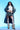A woman stands confidently against a blue background, wearing the LIV FUR TRIM DENIM TRENCH and a white corset top. Her plaid mini skirt pairs elegantly with white lace stockings and fluffy boots. She has curly hair and completes the look with futuristic sunglasses.