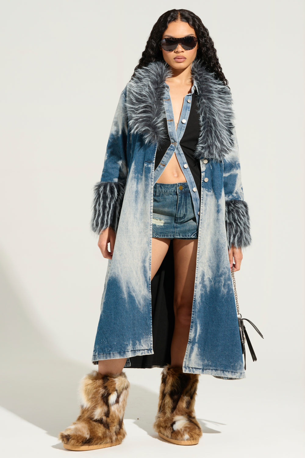 A person dressed in the LIV FUR TRIM DENIM TRENCH pairs it with denim shorts and a black top. They confidently accessorize with dark sunglasses, a handbag, and complement their look with fur boots.
