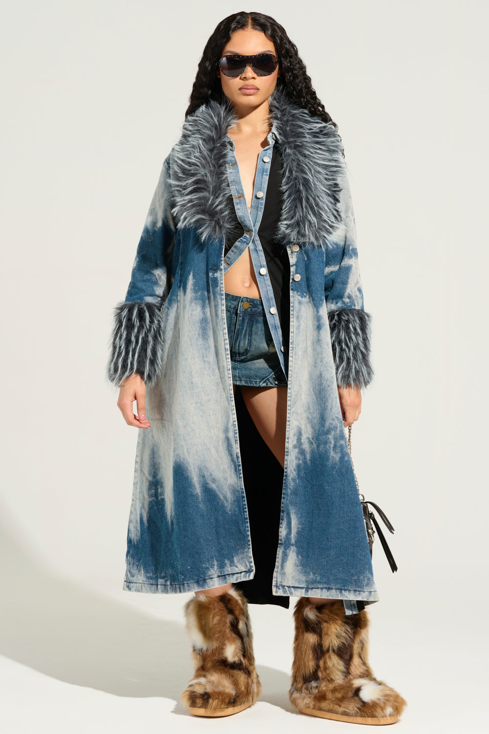 A person sporting the LIV FUR TRIM DENIM TRENCH, complemented by stylish sunglasses, showcases a chic look with a denim trench featuring a fur-trim collar and cuffs. They pair it elegantly with denim shorts, a black top, and furry boots. The simple backdrop accentuates this fashionable ensemble beautifully.