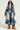 A person sporting the LIV FUR TRIM DENIM TRENCH, complemented by stylish sunglasses, showcases a chic look with a denim trench featuring a fur-trim collar and cuffs. They pair it elegantly with denim shorts, a black top, and furry boots. The simple backdrop accentuates this fashionable ensemble beautifully.