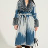 A person confidently stands wearing the LIV FUR TRIM DENIM TRENCH, featuring a chic fur trim collar and cuffs. They have long, curly hair and sport trendy sunglasses. Their outfit is completed with fur boots, and they carry a small bag.