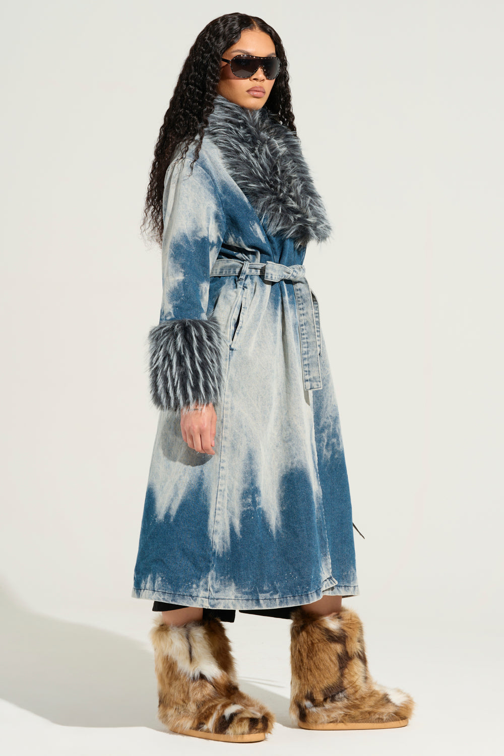 A person in the LIV FUR TRIM DENIM TRENCH, featuring a gray fur trim on the collar and cuffs, is paired with large brown fur boots. They have long curly hair and are wearing sunglasses, standing against a plain background.