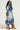 A person in the LIV FUR TRIM DENIM TRENCH, featuring a gray fur trim on the collar and cuffs, is paired with large brown fur boots. They have long curly hair and are wearing sunglasses, standing against a plain background.