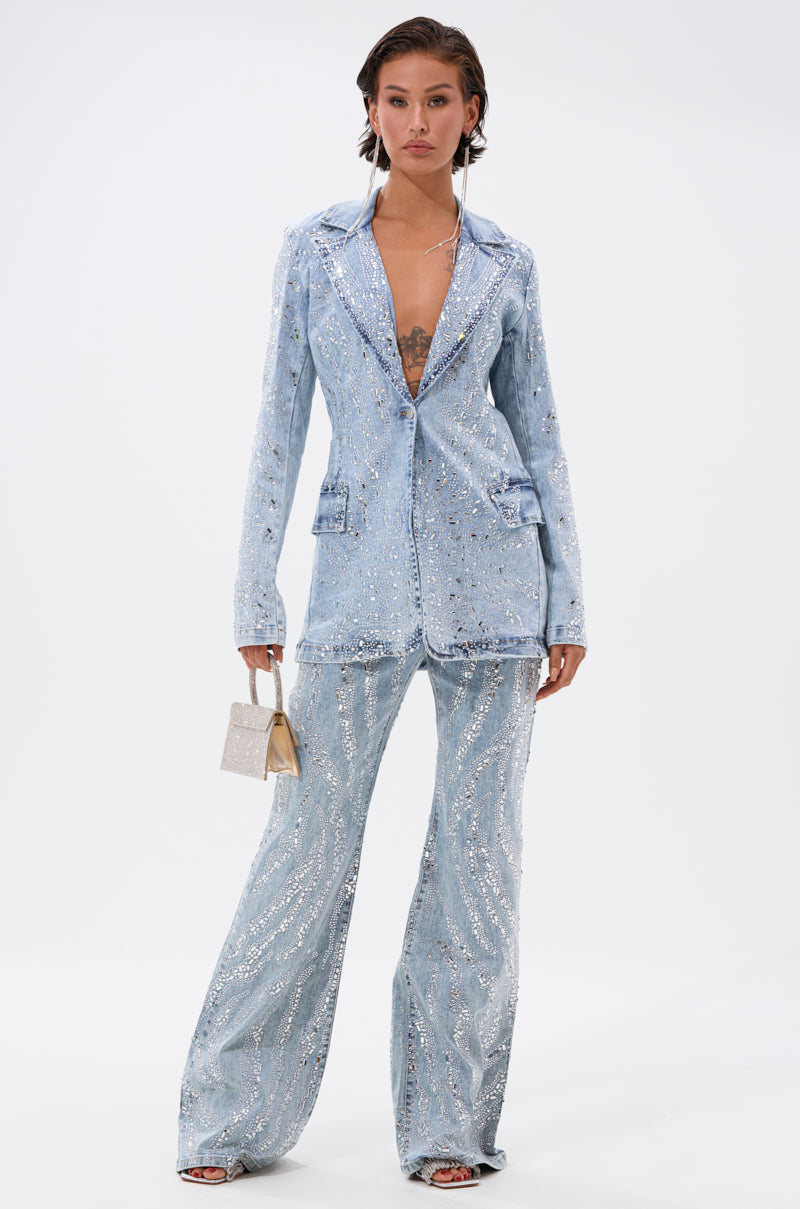 A woman exudes confidence in DIAMONDS DANCING DENIM FLARE PANT—a sparkly light blue flare pant suit with a deep neckline. She pairs it with a metallic purse and slicked-back hair, styled against a crisp white background.