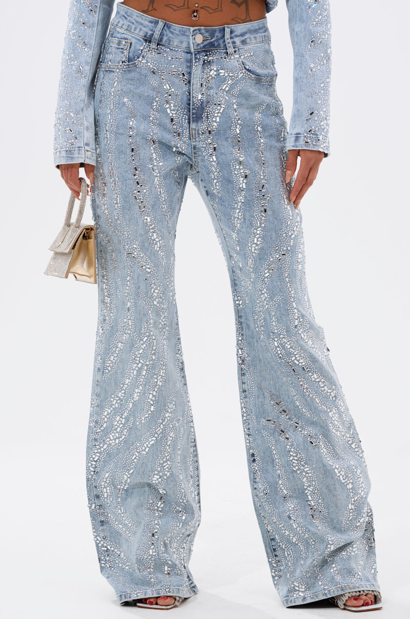 A person wears the DIAMONDS DANCING DENIM FLARE PANT—light blue, high-waisted jeans adorned with silver sequin waves—paired with a metallic handbag and open-toe shoes.