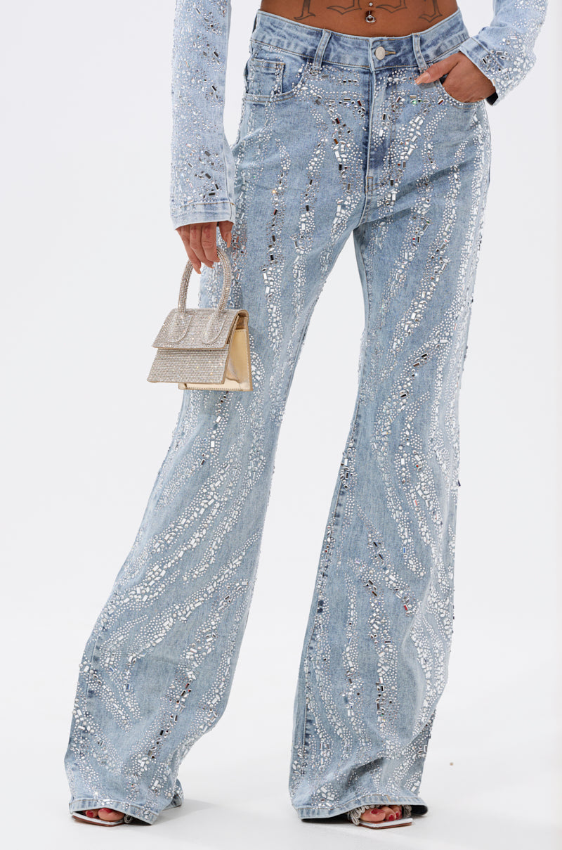 A person wears DIAMONDS DANCING DENIM FLARE PANT—high-waisted, light-wash jeans with jeweled, wavy embellishments and a relaxed flared fit—styled with a sparkly handbag and open-toe heels.