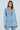 Clad in the DIAMONDS DANCING DENIM BLAZER, a person teams this light blue gem-embellished denim blazer with matching pants. With straight brown hair, they pose against a light blue background, exuding effortless luxury.