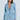 A woman with long brown hair is wearing the DIAMONDS DANCING DENIM BLAZER, a luxe light blue sequined jacket with matching pants and deep V-neck/button closure. She stands against a light blue background, looking right, with sparkling silver jewels dazzling as her earrings.