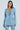 A woman with long brown hair is wearing the DIAMONDS DANCING DENIM BLAZER, a luxe light blue sequined jacket with matching pants and deep V-neck/button closure. She stands against a light blue background, looking right, with sparkling silver jewels dazzling as her earrings.