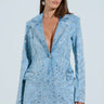 A woman with long brown hair is wearing the DIAMONDS DANCING DENIM BLAZER, a luxe light blue sequined jacket with matching pants and deep V-neck/button closure. She stands against a light blue background, looking right, with sparkling silver jewels dazzling as her earrings.
