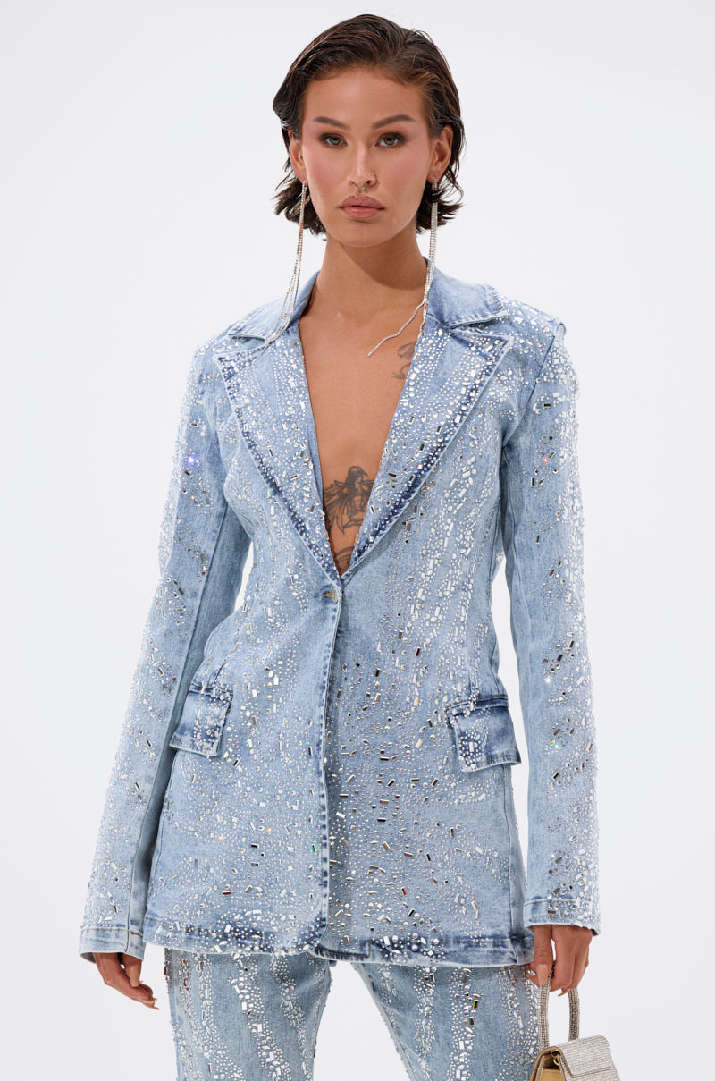 Wearing the DIAMONDS DANCING DENIM BLAZER and matching pants, a person with short hair styles the light blue, sequin-embellished blazer buttoned over bare skin, showcasing a chest tattoo and carrying a small silver purse.