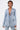 Wearing the DIAMONDS DANCING DENIM BLAZER and matching pants, a person with short hair styles the light blue, sequin-embellished blazer buttoned over bare skin, showcasing a chest tattoo and carrying a small silver purse.