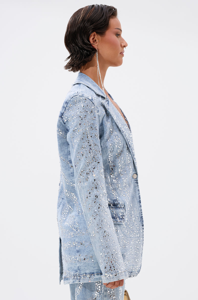 A person with short, dark hair models the DIAMONDS DANCING DENIM BLAZER, an oversized blazer adorned with silver sequins and embellishments, standing in profile against a plain white background.