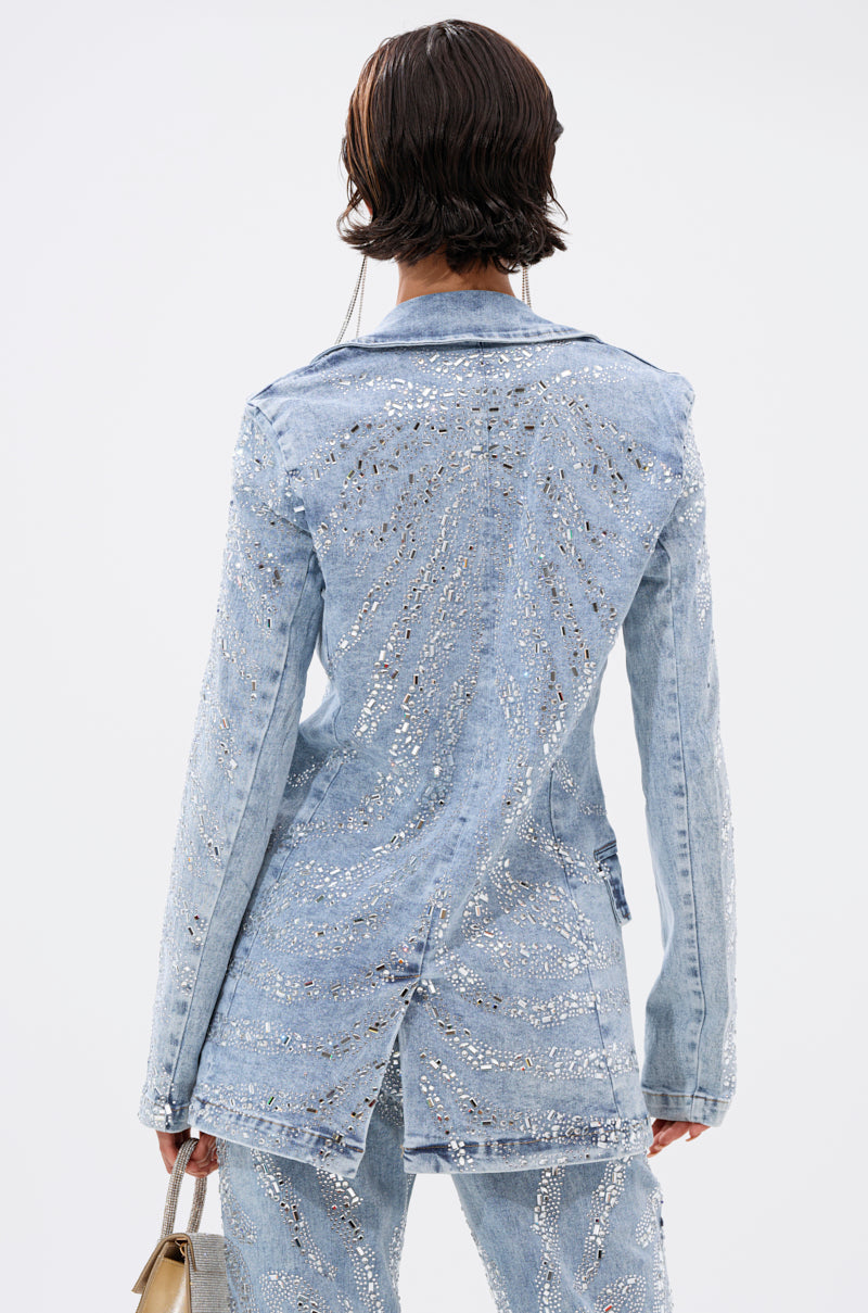 A person with short, wet-look hair is seen from the back wearing the DIAMONDS DANCING DENIM BLAZER, adorned with sparkling silver sequins. They hold a small metallic handbag, highlighting this dazzling statement piece.
