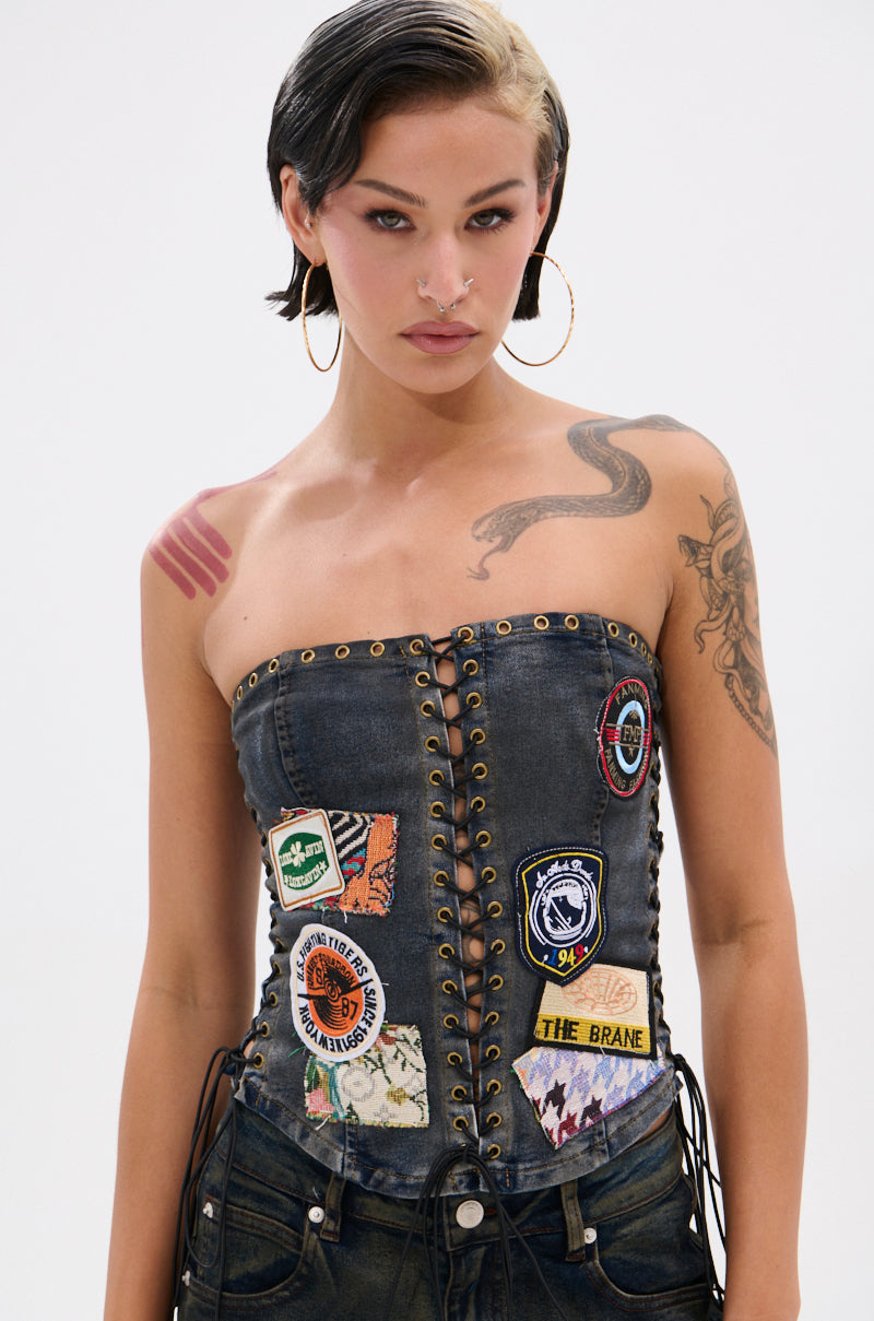 A person with short dark hair and a blonde streak wears the ARTS AND CRAFTS PATCH WORK DENIM CORSET, which is strapless with lace-up panels and vibrant patchwork. They style it with hoop earrings, bold makeup, and display tattoos on their arm and shoulder.