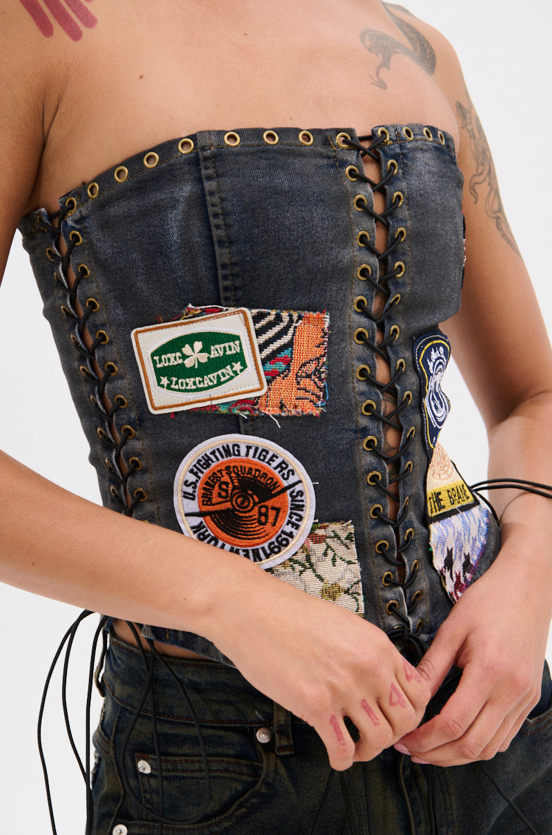Wearing the ARTS AND CRAFTS PATCH WORK DENIM CORSET, a person adjusts its front laces, revealing tattoos through the dark denim with lace-up panels and vibrant embroidered patches.