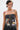 A person with short dark hair and a blonde streak wears the ARTS AND CRAFTS PATCH WORK DENIM CORSET, which is strapless with lace-up panels and vibrant patchwork. They style it with hoop earrings, bold makeup, and display tattoos on their arm and shoulder.