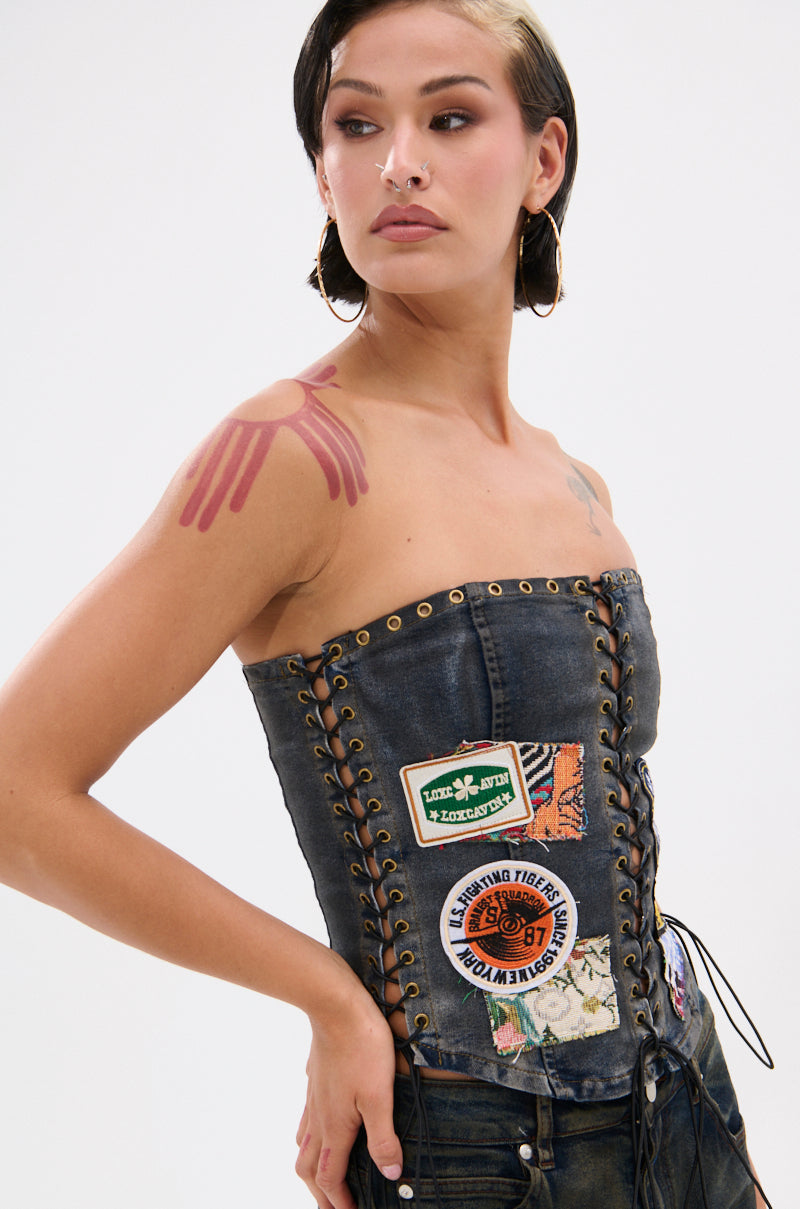 A person with hoop earrings and face piercings wears the ARTS AND CRAFTS PATCH WORK DENIM CORSET, featuring lace-up panels and colorful patches, with a red painted shoulder design, posing against a plain background.