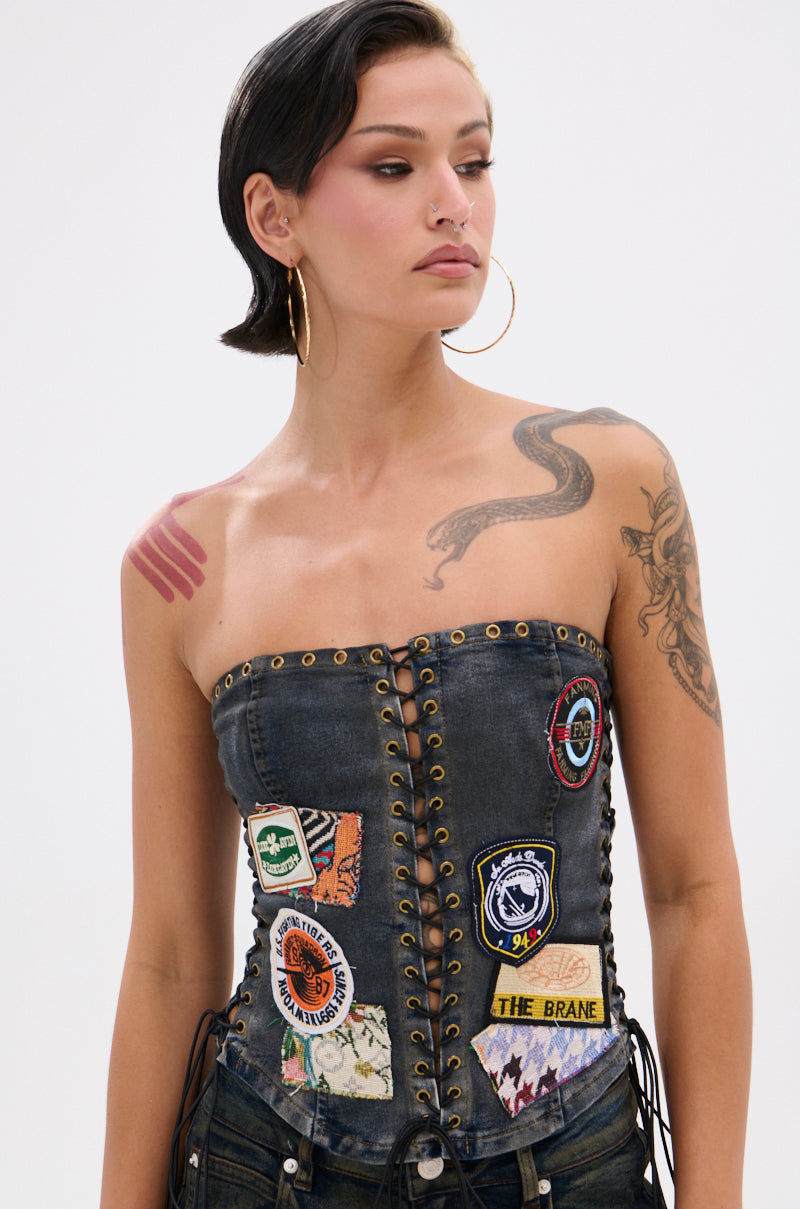 A woman with short dark hair, hoop earrings, and visible tattoos models the ARTS AND CRAFTS PATCH WORK DENIM CORSET featuring lace-up panels and vibrant patchwork. She looks to the side against a plain background.