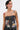 A woman with short dark hair, hoop earrings, and visible tattoos models the ARTS AND CRAFTS PATCH WORK DENIM CORSET featuring lace-up panels and vibrant patchwork. She looks to the side against a plain background.