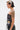 A person with short, black and platinum-blonde hair and a Medusa tattoo on their upper arm wears the ARTS AND CRAFTS PATCH WORK DENIM CORSET with lace-up panels and embroidered patches, paired with denim pants and hoop earrings.