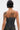 Seen from the back, a person with short, dark hair and shoulder tattoos—including a woman's face and a snake—wears the ARTS AND CRAFTS PATCH WORK DENIM CORSET with lace-up panels and jeans. A pink handprint appears on their right shoulder.