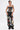 Confidently posed against a white background, a person with short dark hair, tattoos, and black platform shoes models the ARTS AND CRAFTS PATCH WORK DENIM CORSET with lace-up panels and loose pants featuring colorful embroidered patches.
