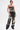 A woman poses confidently in the ARTS AND CRAFTS PATCH WORK DENIM CORSET—strapless with lace-up panels—paired with loose pants, both featuring colorful embroidered patches and graphic designs. One hand is in her pocket; her hair is wet and slicked back.