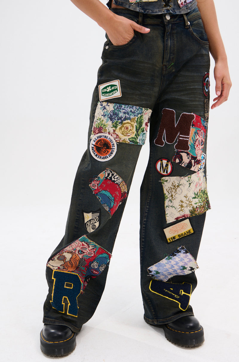 Wearing ARTS AND CRAFTS PATCH WORK DENIM PANTS featuring vintage dark-wash denim, colorful patches, embroidered letters, floral prints, and badges; only the unique lower half is shown against a plain white background.