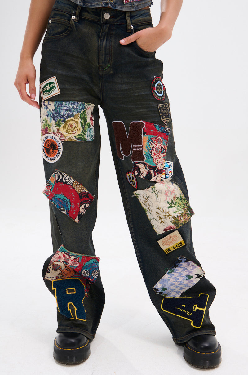 The lower half of a person wearing ARTS AND CRAFTS PATCH WORK DENIM PANTS—vintage dark-wash denim pants with colorful fabric patches and letter appliqués—paired with black boots is shown against a plain background.