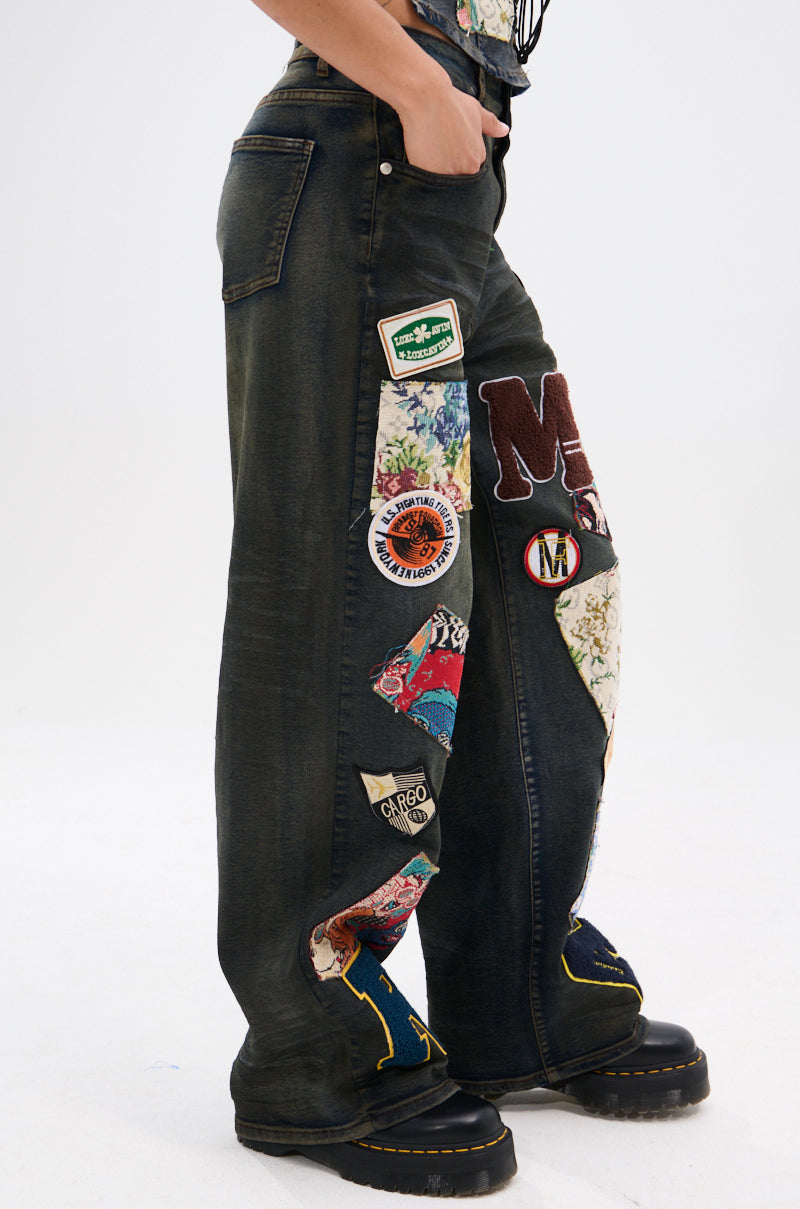 A person wears ARTS AND CRAFTS PATCH WORK DENIM PANTS, featuring colorful patches and embroidery, with black platform shoes. The photo showcases the pants from waist to shoes against a plain background.