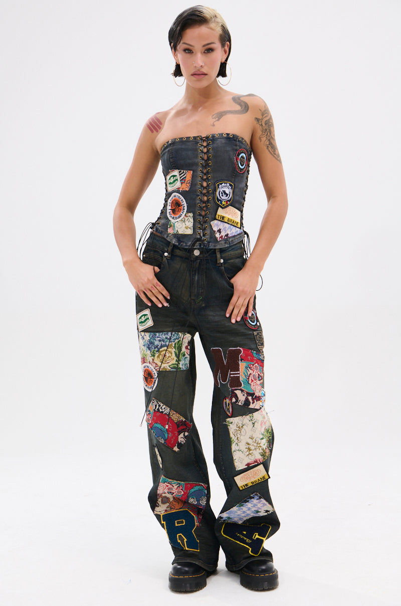A person with short dark hair and tattoos wears ARTS AND CRAFTS PATCH WORK DENIM PANTS with colorful patches, styled with a strapless corset top, standing confidently against a plain white background.