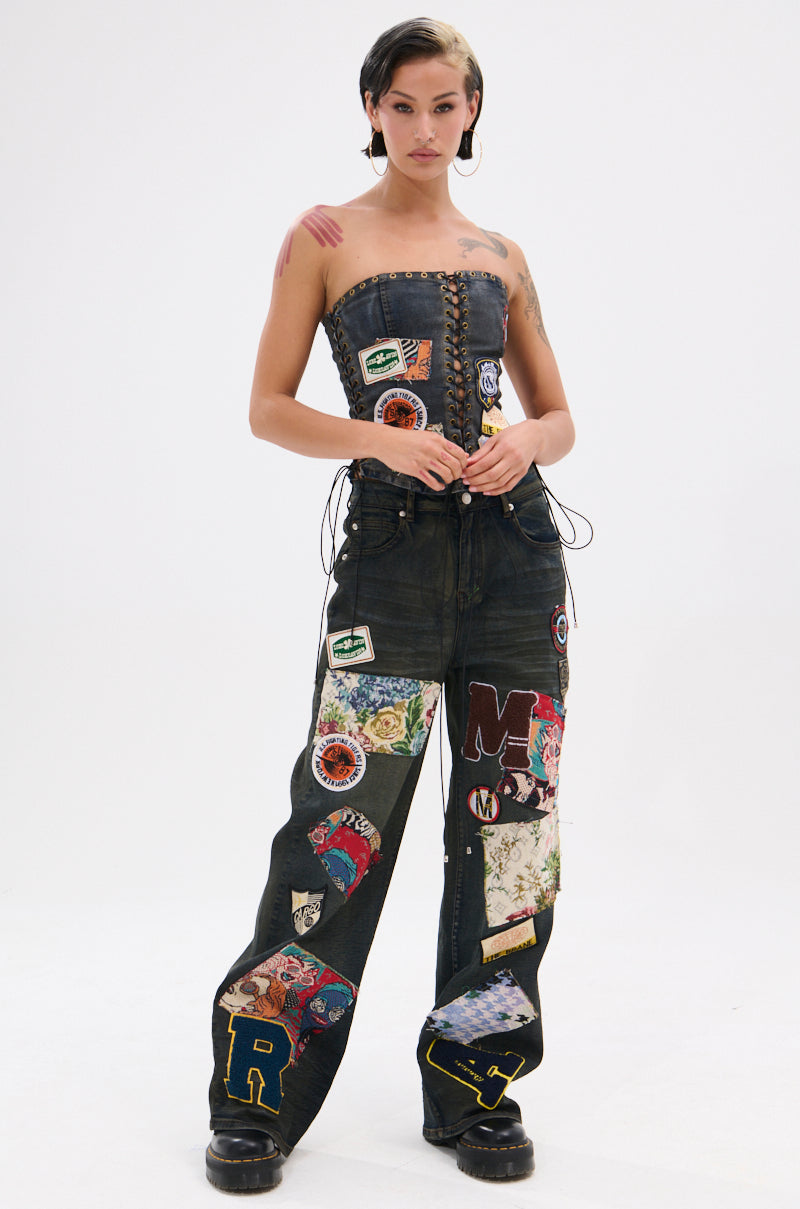 A woman wears ARTS AND CRAFTS PATCH WORK DENIM PANTS featuring vintage dark-wash patches and colorful graphics, styled with a strapless lace-up corset, black shoes, short dark hair, and visible arm and shoulder tattoos.