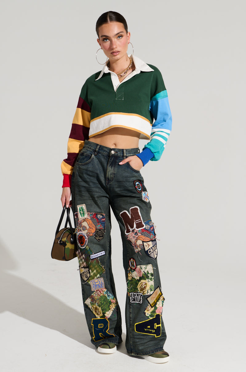 A woman is posing against a plain background, wearing a cropped rugby-style shirt with multicolored stripes and the FOREVER DENIM PANT adorned with assorted patches. She is holding a small camouflage-patterned handbag, has her hair tied back, and is wearing large hoop earrings.