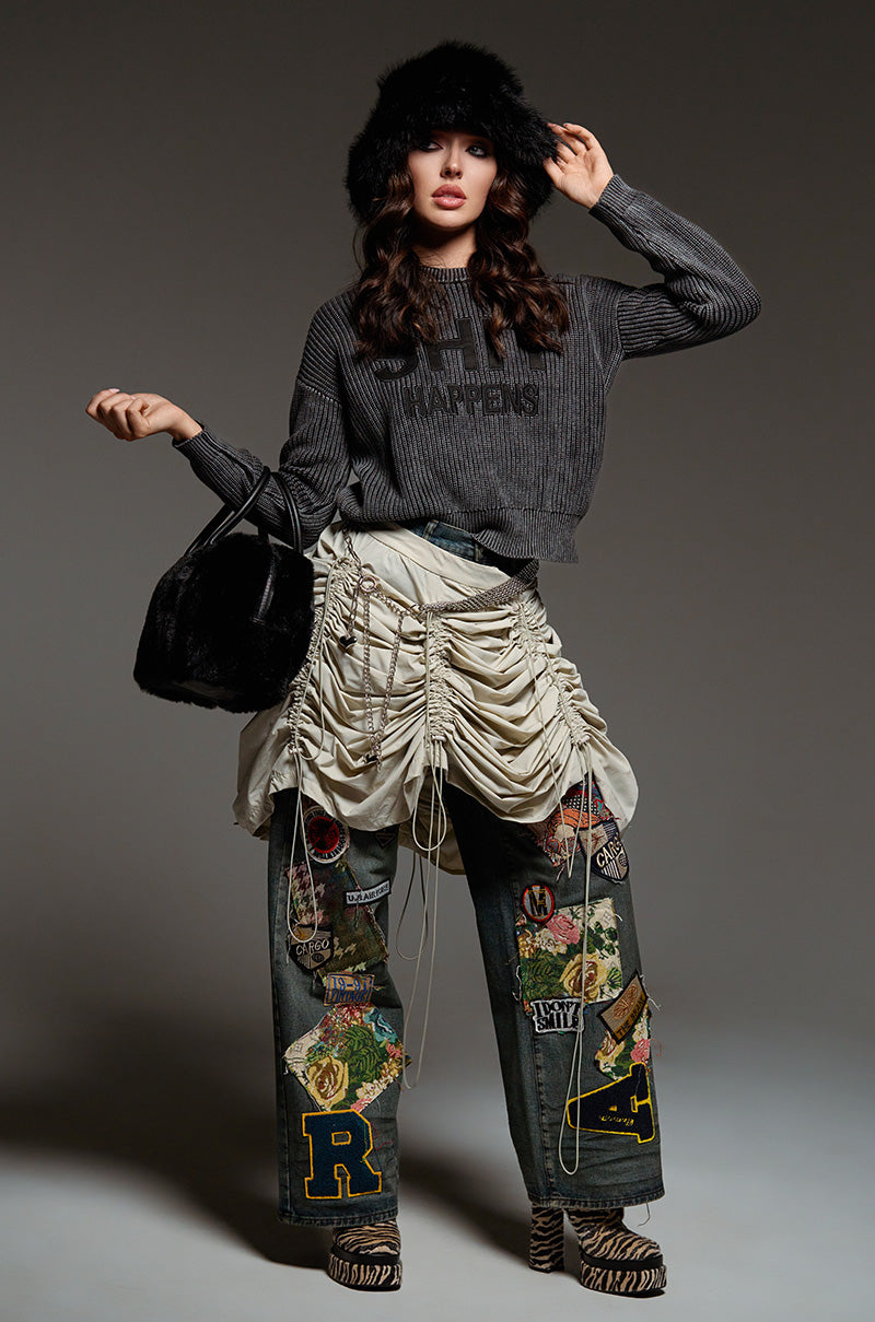 A woman poses stylishly, wearing a dark hat, a striped sweatshirt, and the FOREVER DENIM PANT featuring elaborate ties and embroidered designs. She carries a black bag and stands against a neutral background.