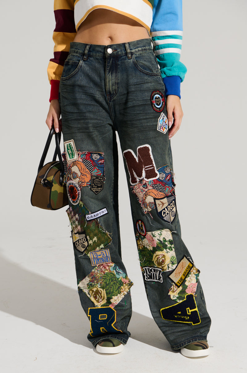 A person stands modeling the FOREVER DENIM PANT, featuring a high-waisted, wide-leg silhouette adorned with various colorful patches and logos. They are also wearing a cropped, multicolored, long-sleeve top and holding a small camouflage-patterned handbag. Only part of their head is visible in the image.