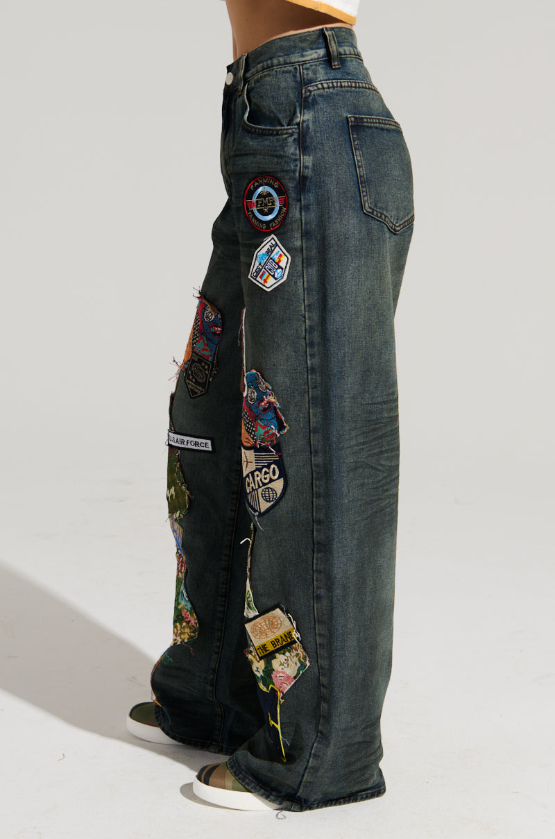 A person is wearing the FOREVER DENIM PANT, a pair of dark, high-rise jeans with a wide-leg silhouette decorated with colorful patches and intricate embroidery. These jeans feature a side patch pocket and have frayed edges on some of the patches. The person completes the look with white sneakers featuring a black stripe.