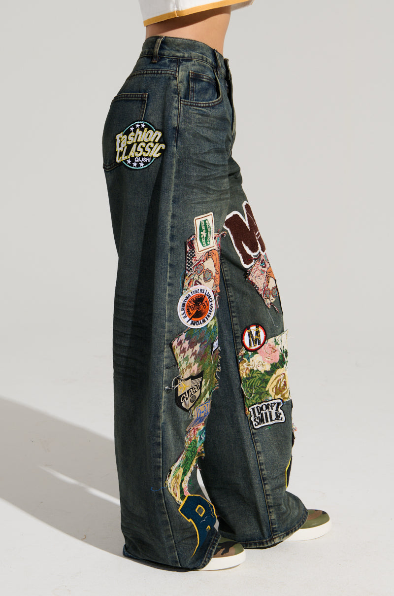 A person is wearing the FOREVER DENIM PANT, featuring high-rise, dark blue wide-leg denim adorned with various colorful patches and embroidery such as "Fashion Classic" and a smiley face. These embroidered jeans have a distinct, eclectic style with a mix of graphics and textures.