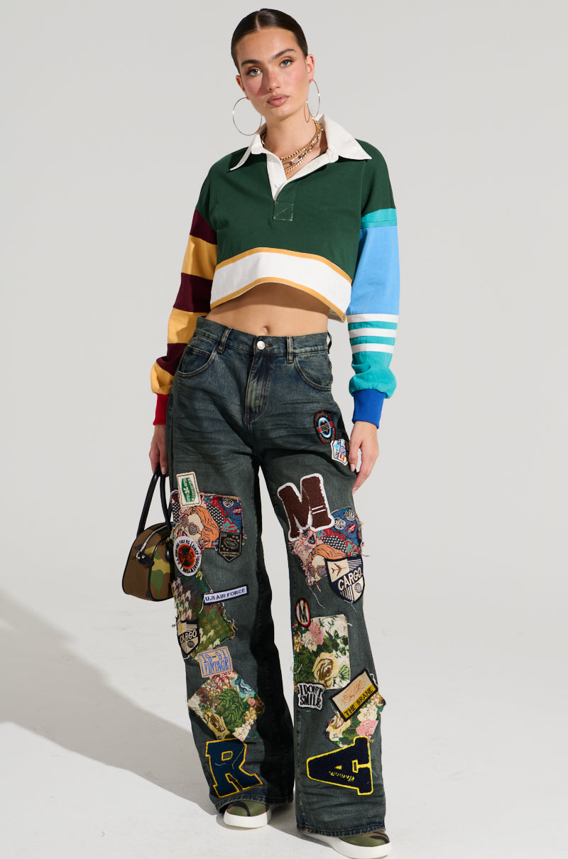 A woman with slicked-back hair is wearing a cropped, color-blocked rugby shirt and FOREVER DENIM PANT high-rise jeans decorated with various colorful patches. She accessorizes with large hoop earrings, layered necklaces, and a small handbag while standing against a plain background.