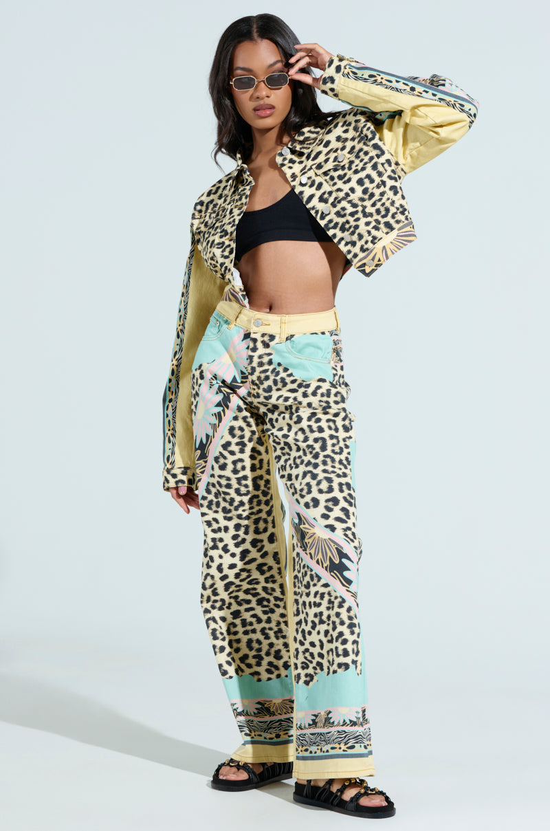 The woman poses in Trust Your Instincts wide-leg denim, featuring a leopard-print pattern with long sleeves and accented in light turquoise. She accessorizes with dark sunglasses, a black top, and sandals against a light-colored background.