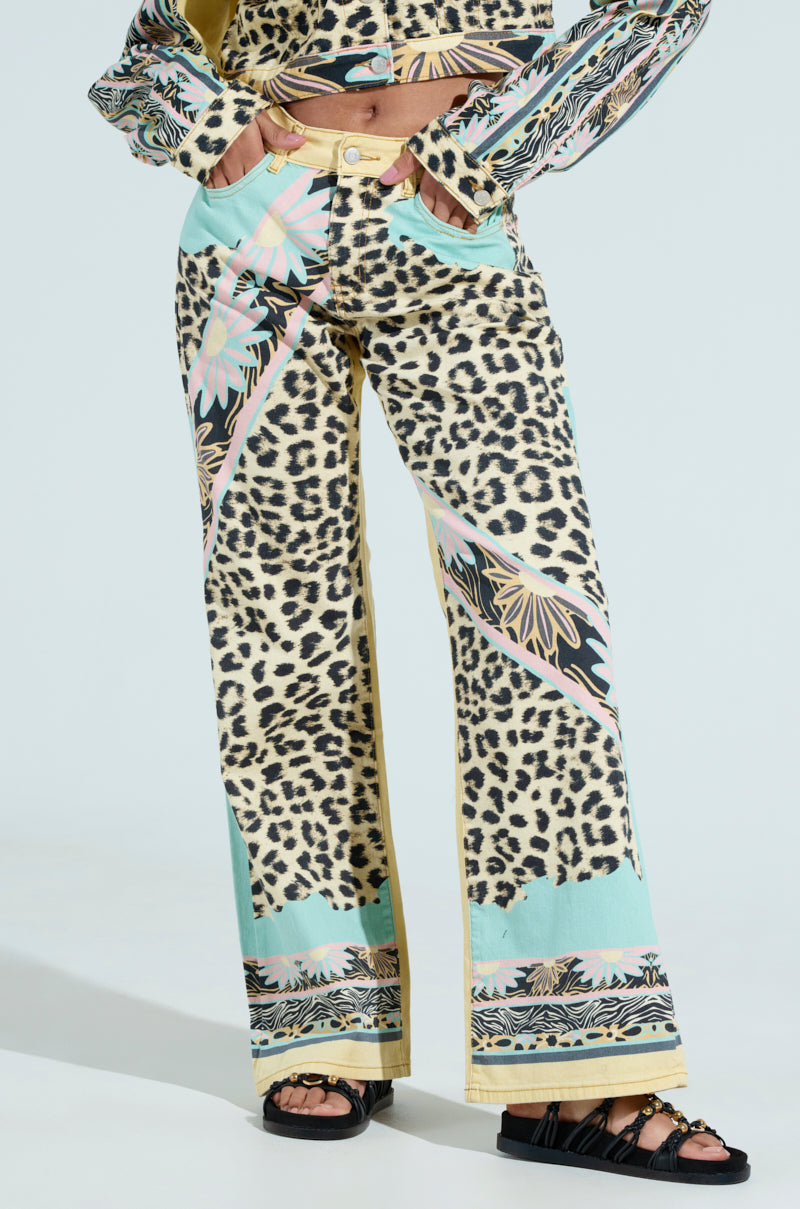 Someone wearing TRUST YOUR INSTINCTS WIDE LEG DENIM, featuring vibrant leopard-print patterns and pastel abstract designs, paired with black sandals. A solid light background highlights the striking design of the clothing.