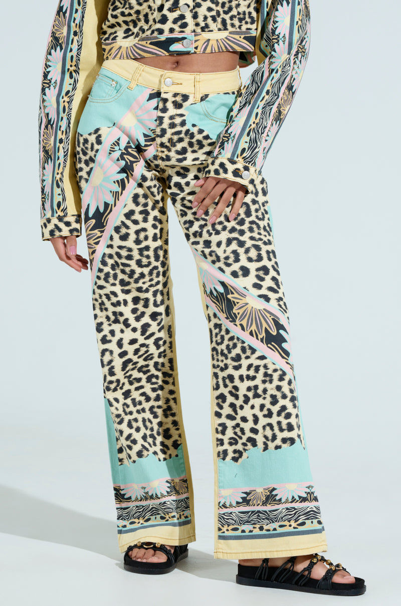 A person wears the TRUST YOUR INSTINCTS WIDE LEG DENIM—a colorful leopard-print pant with pastel florals and geometric patterns. The high-rise design adds a unique twist, paired with a matching top and black sandals, all set against a light gray background.