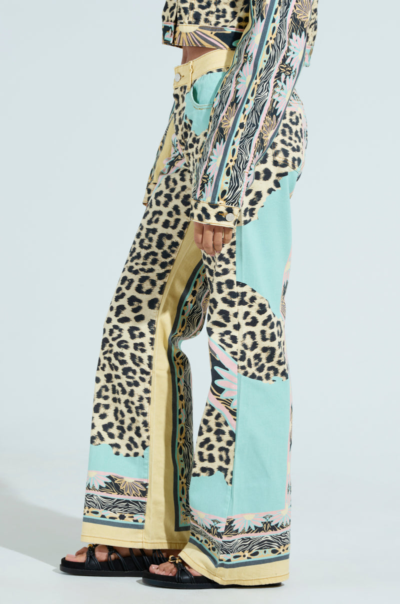 Someone is wearing the TRUST YOUR INSTINCTS WIDE LEG DENIM, featuring a high-rise and wide-leg patchwork design with leopard print, pastel blue, and yellow sections. They complement these intricate pants with black sandals against a plain light background.