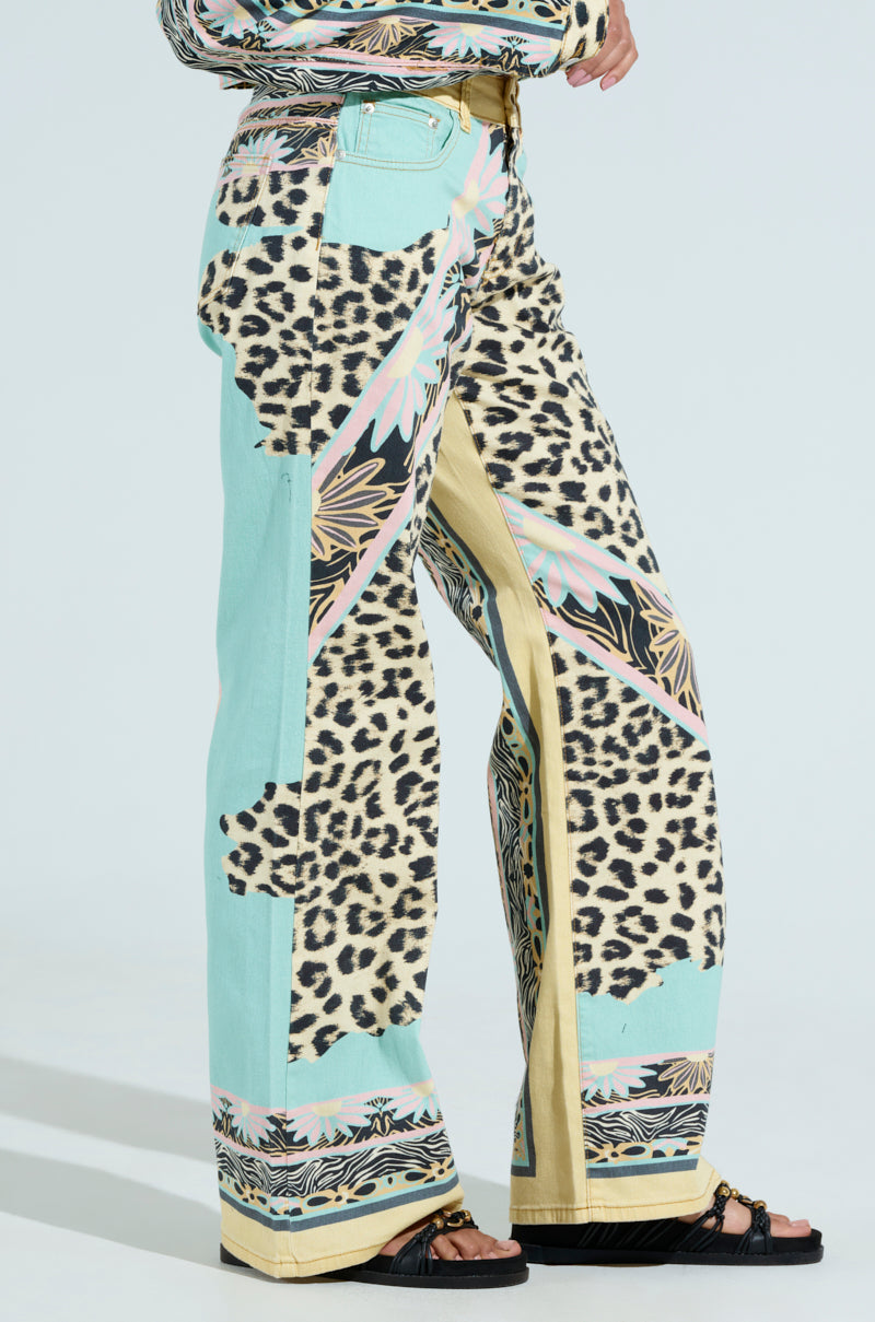 A person wears the "TRUST YOUR INSTINCTS WIDE LEG DENIM," featuring a colorful leopard-print design with geometric patterns, light blue, and yellow accents. These high-rise pants are paired with black sandals against a plain light background.