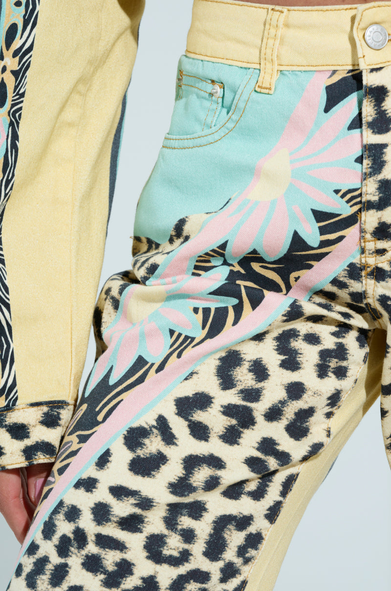 Close-up of TRUST YOUR INSTINCTS WIDE LEG DENIM featuring a vibrant floral and leopard-print in pastel shades. The design features large flowers and animal spots, highlighted by a yellow waistband and pocket, with a hand partially visible.