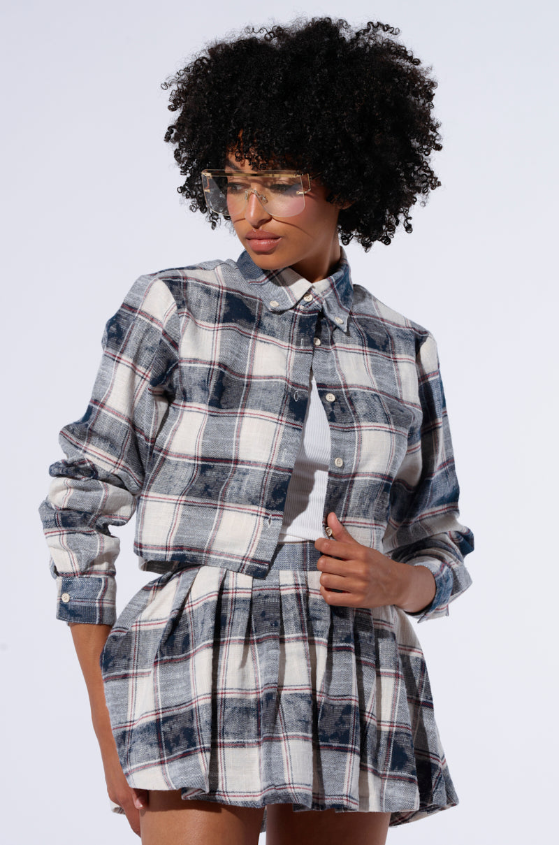 A person with curly hair and glasses models a 100% cotton plaid outfit consisting of a BELLA PLAID BUTTON DOWN SHIRT and a Bella Plaid Pleated Mini Skirt. They are standing against a plain white background, touching their shirt with one hand. 0